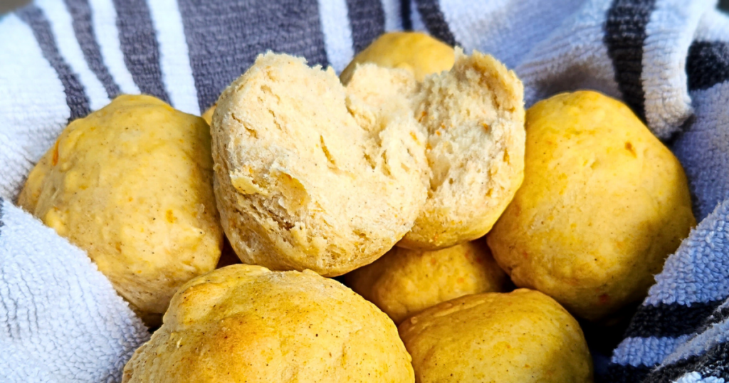 A close-up of several freshly baked, golden-brown dinner rolls in a basket lined with a white and blue striped cloth. One roll is broken open, revealing its soft, fluffy interior.