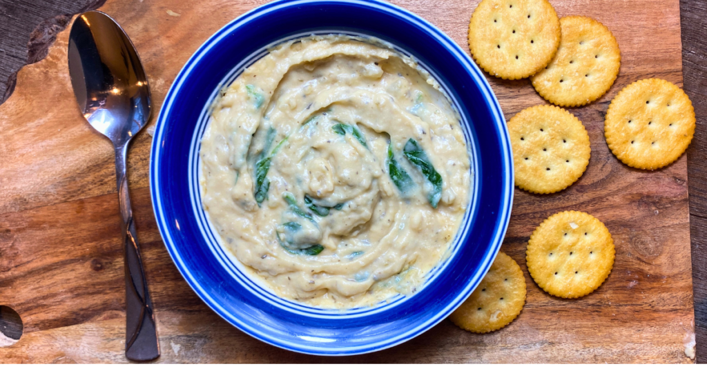 A blue bowl filled with creamy spinach dip sits on a wooden cutting board. The dip has hints of green spinach leaves swirled throughout. A spoon is placed to the left of the bowl, and a few round Ritz crackers are arranged to the right.