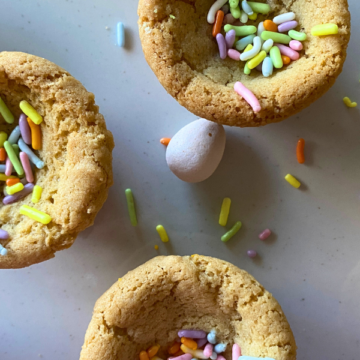 A buttery, chewy sugar cookie cup is filled with a colorful candy coated chocolate egg, and topped with festive sprinkles.